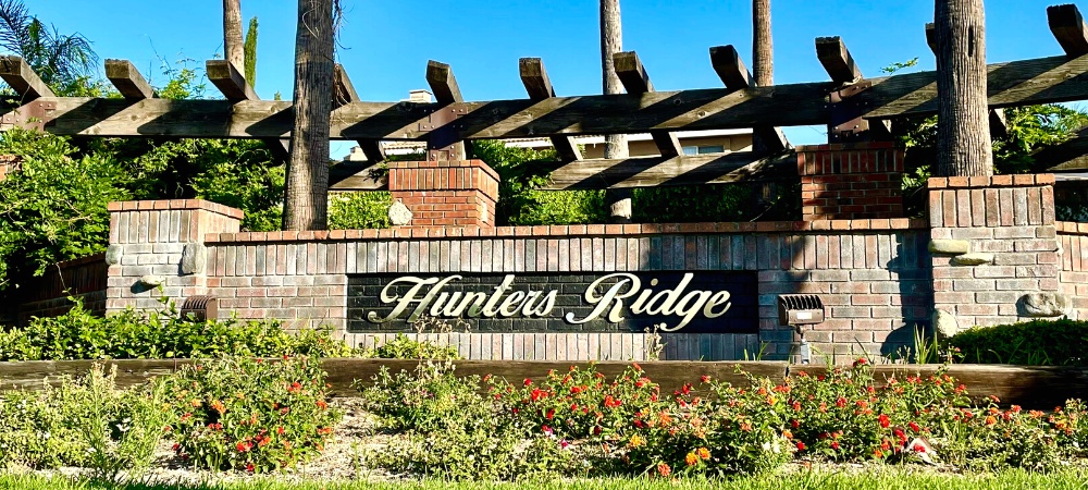 Hunters Ridge Signage attached to a brick wall surrounded by greenery and colorful flowering plants-Hunters Ridge Homes For Sale-Featured Image-Alvin Tapia
