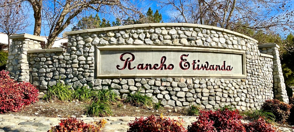 Stone wall with Rancho Etiwanda name with red font color tall trees in the background-Rancho Etiwanda Homes For Sale-Featured Image-Alvin Tapia